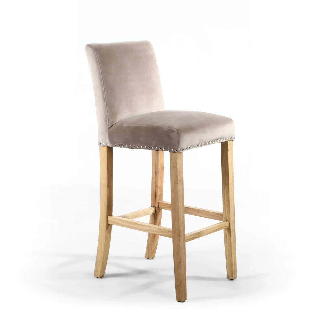 A beige velvet bar stool with a cushioned backrest and seat, adorned with silver studs. It features light wooden legs and footrests, exuding elegance and comfort.